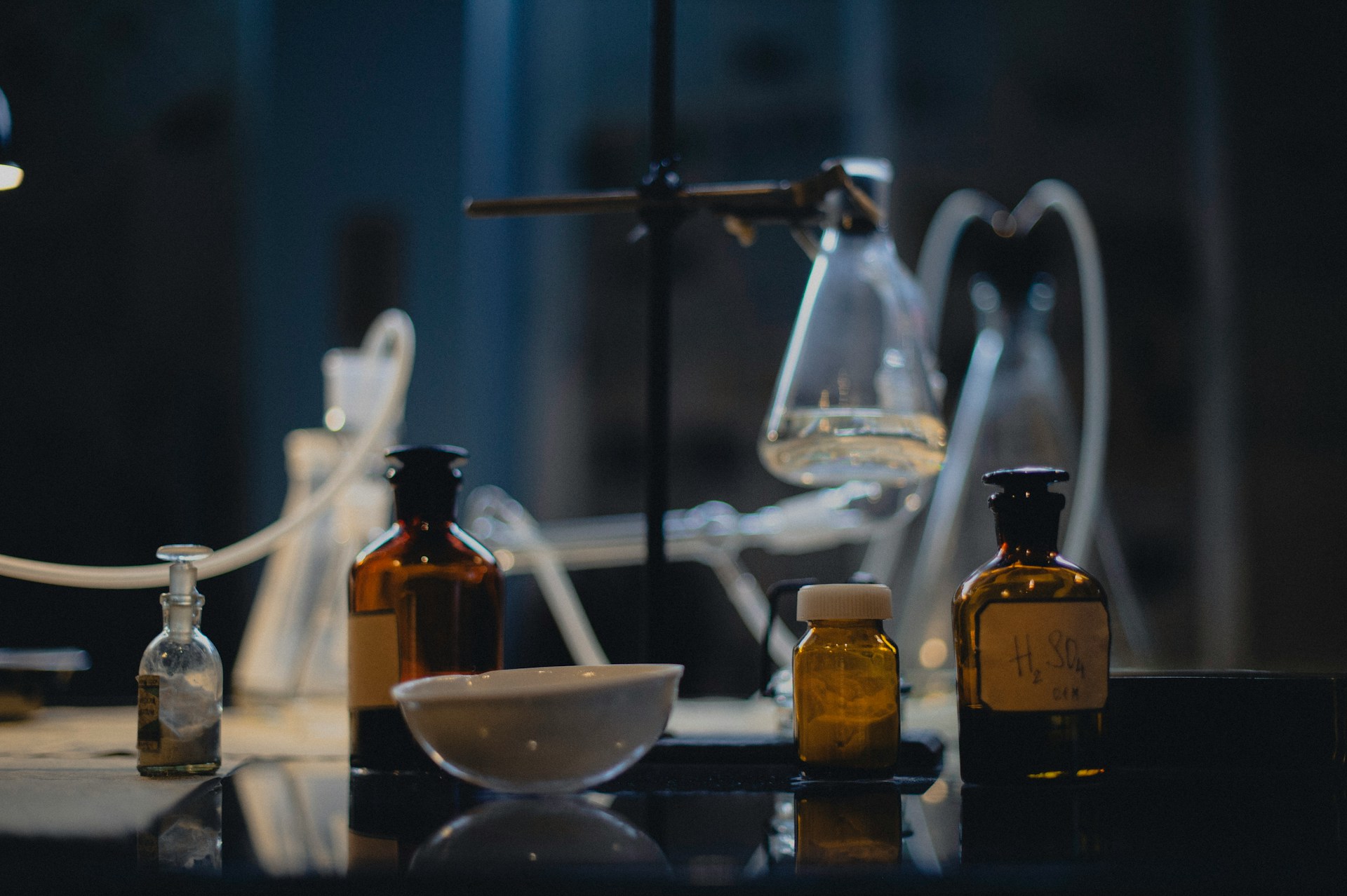 Laboratory glassware in low light: a flask on a stand, a white evaporating dish and several amber reagent bottles