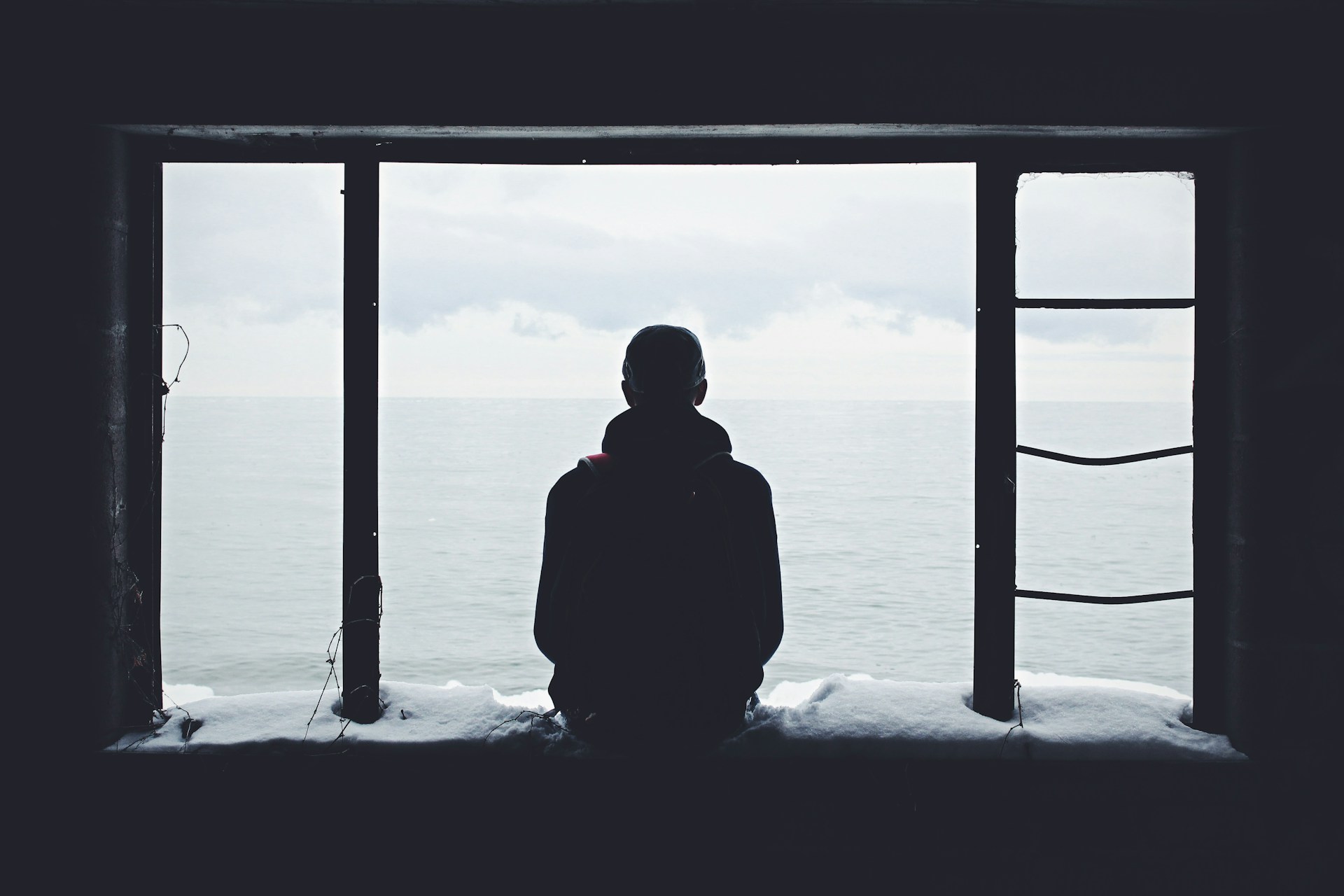 A figure seen from behind, sitting in a large empty window frame, looking out at a flat grey sea