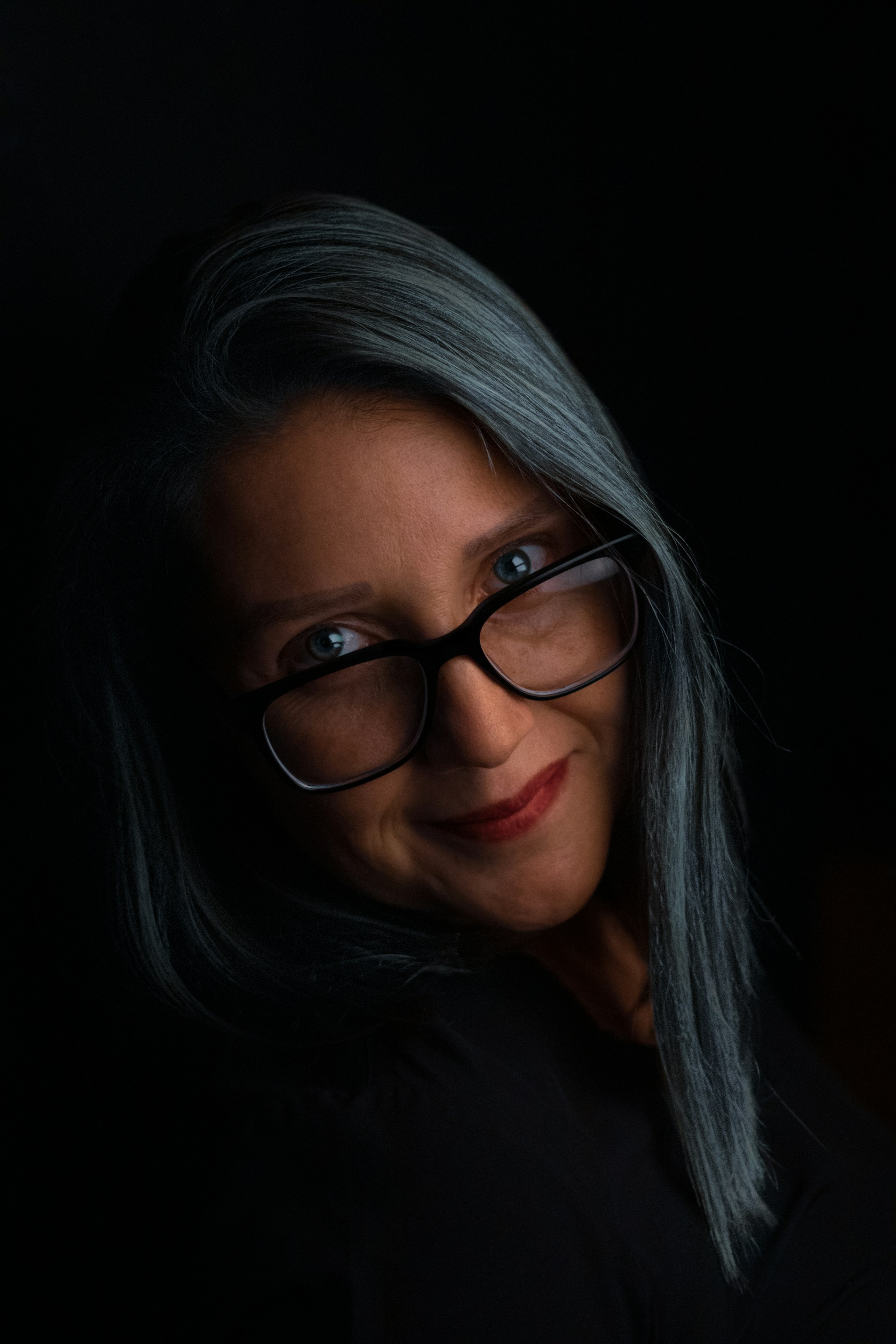 Portrait: a woman with long grey hair and black-framed glasses, lit against a black background