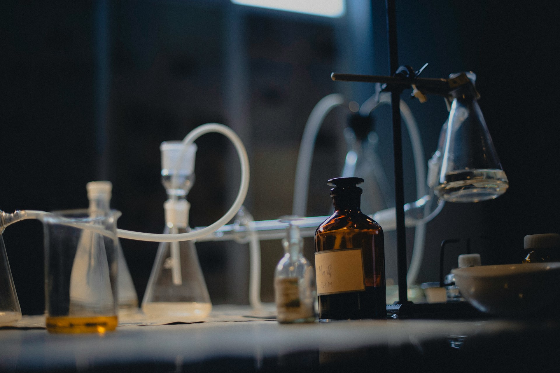 A dimly lit laboratory bench: glass flasks and tubing on a stand, with amber reagent bottles in the foreground