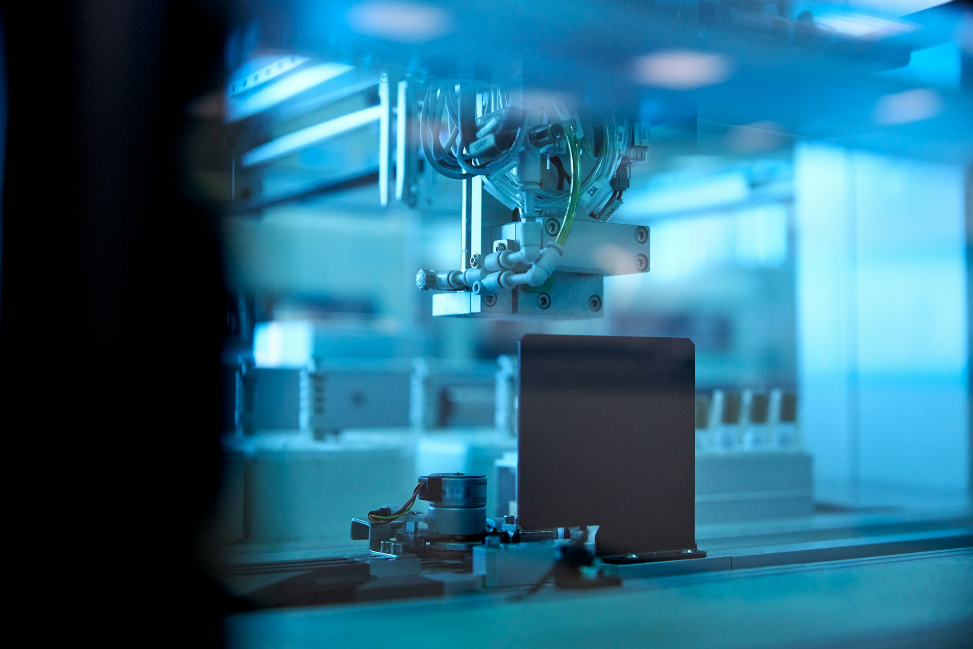 An automated laboratory instrument seen through glass under blue light: a robotic head poised above a row of sample trays