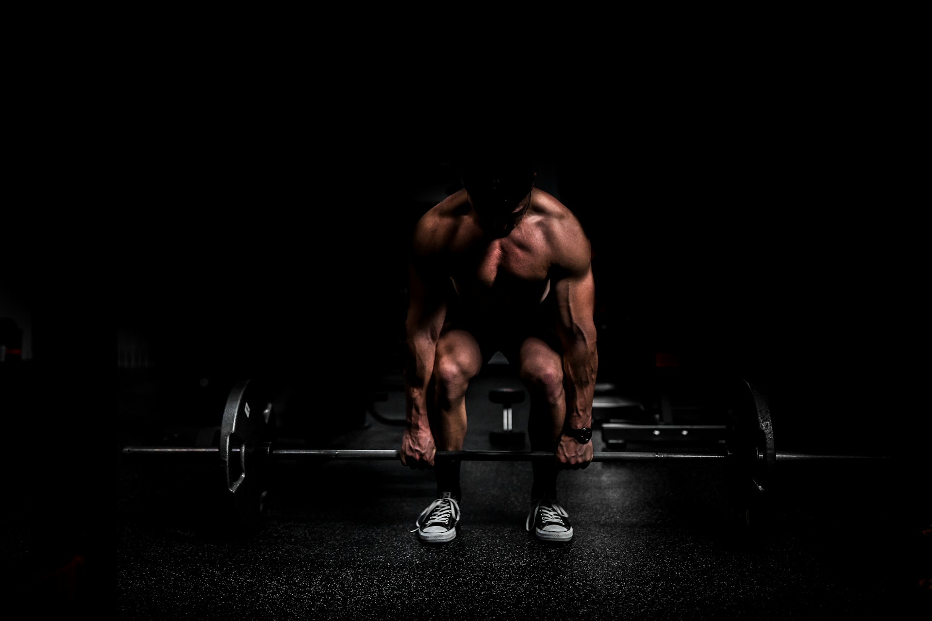 A dark gym: a lifter bent over a loaded barbell, lit from above so that only the shoulders, arms and shoes catch the light